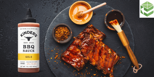Kinders Gold BBQ sauce bottle next to glazed ribs, honey, and spices on a black slate, highlighting rich BBQ flavor.